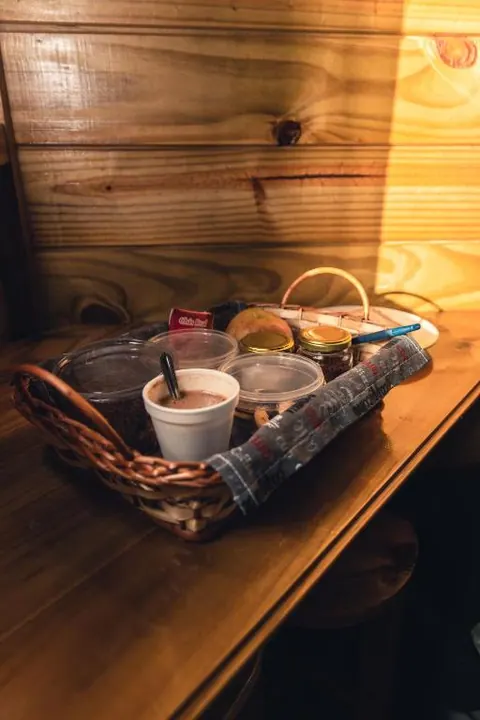 Cesta de café da manhã com bolo, pães, geleias e uma xícara de bebida quente sobre a mesa de madeira
