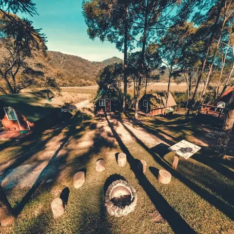 Fim de tarde dourado sobre o gramado, a roda de fogueira com tocos de madeira e os chalés, com a serra ao fundo