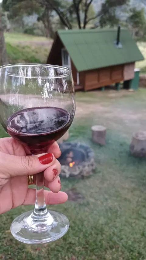 Taça de vinho tinto em primeiro plano e a fogueira acesa no pátio, com um chalé ao fundo