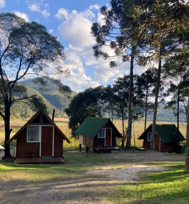 Três chalés de madeira com telhado verde entre araucárias, com a serra ao fundo